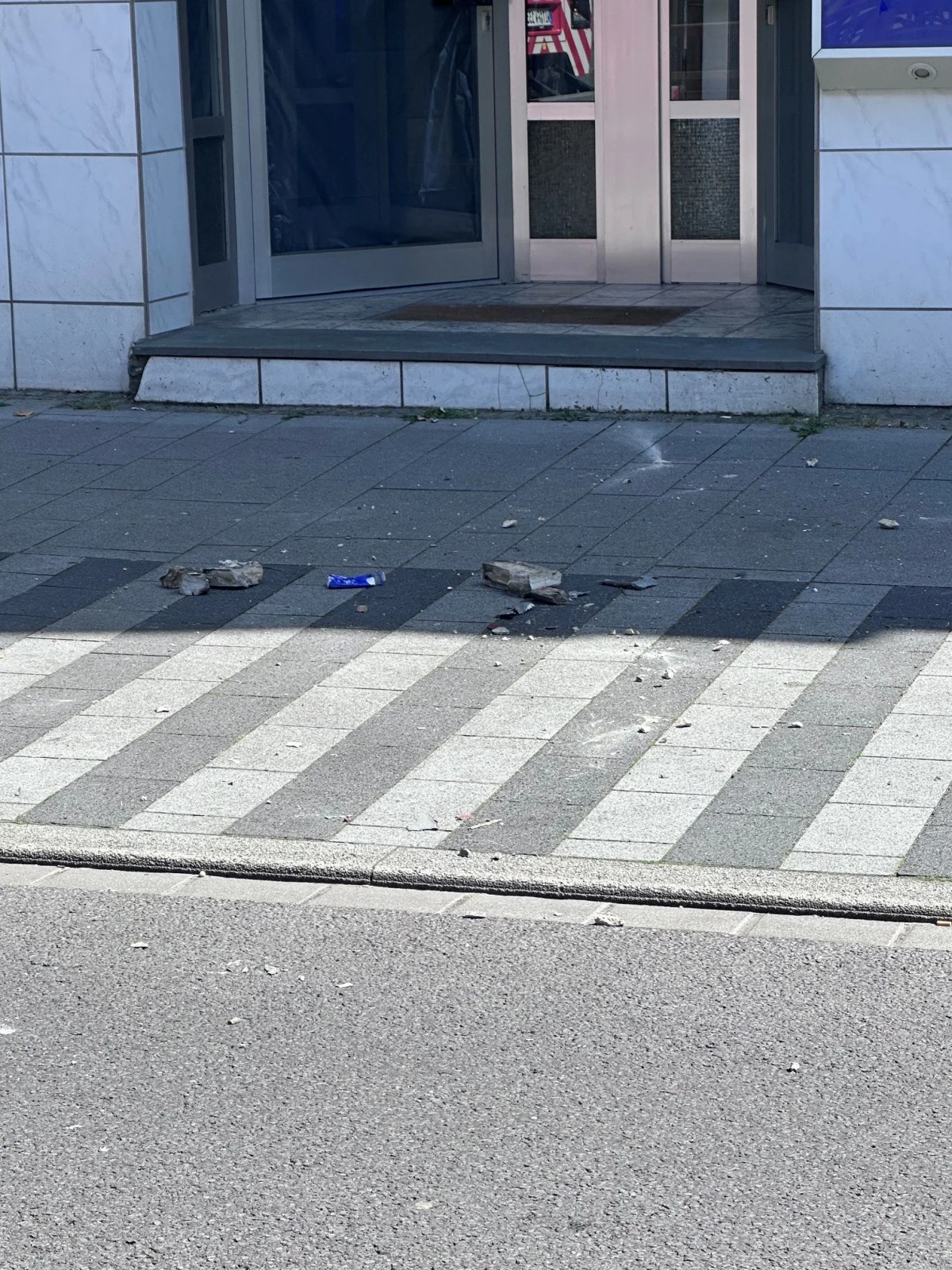 Zerstörte Pflastersteine und Trümmer liegen verstreut auf einem Gehweg vor einem Gebäudeeingang, es ist ein klarer Tag.