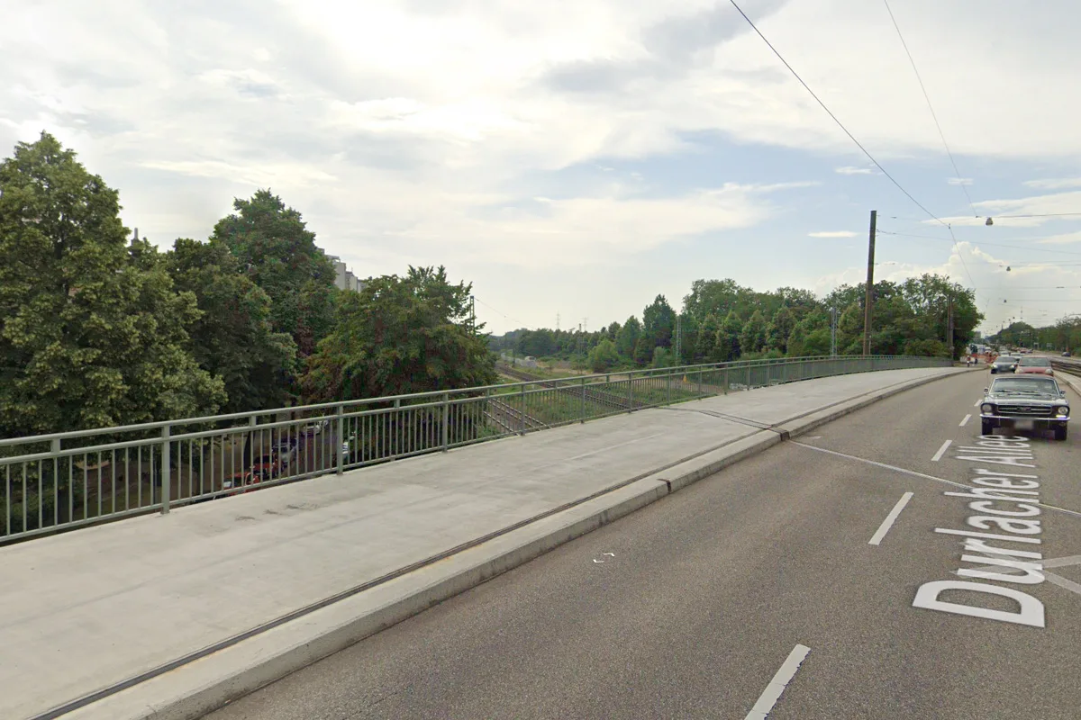 Eine Brücke mit einem Bürgersteig und einigen Autos auf der Straße über Bahngleise. Bäume flankieren die Szenerie bei klarem Himmel.