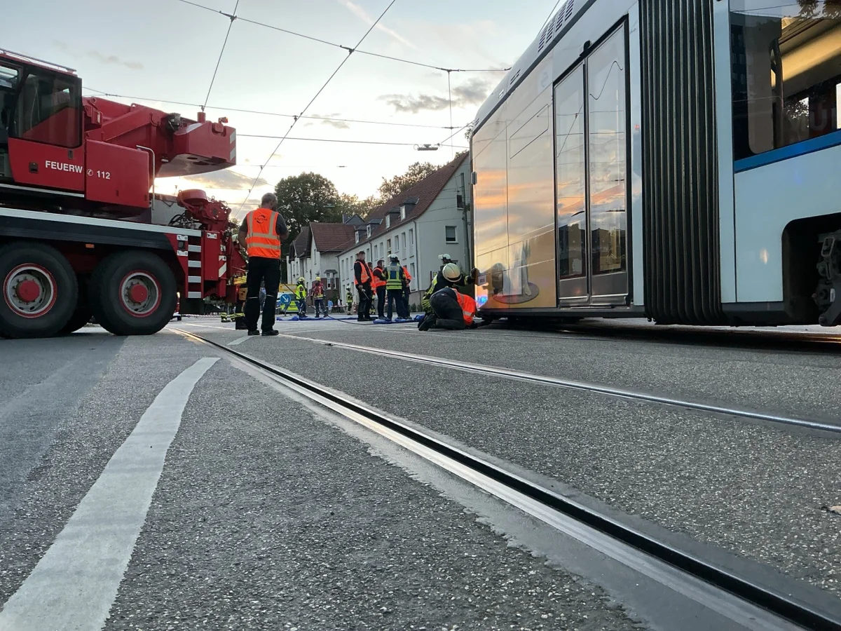 Eine entgleiste Straßenbahn wird von Einsatzkräften und einem Feuerwehrkran auf einer städtischen Straße bearbeitet. Ein Feuerwehrmann in orangefarbener Warnweste kniet neben der Straßenbahn.