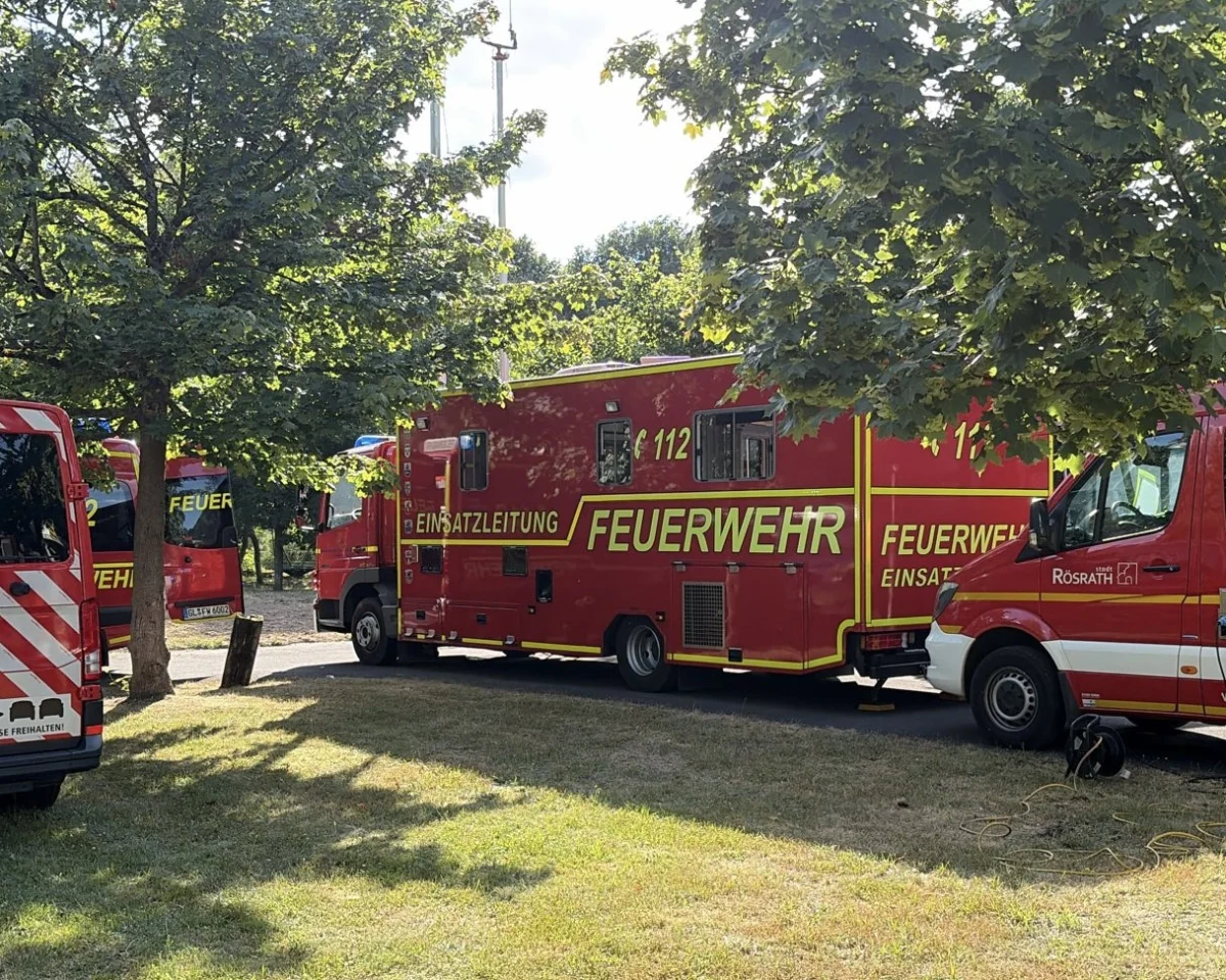 Mehrere rote Feuerwehrfahrzeuge der Einsatzleitung stehen auf einer grasbewachsenen Fläche unter Bäumen bei Tageslicht.