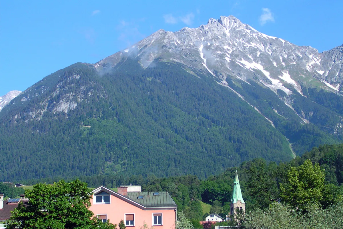 Eine alpine Landschaft mit einem hohen, schneebedeckten Berg, umgeben von dichten Wäldern. Im Vordergrund sind einige Gebäude und eine Kirche zu sehen.
