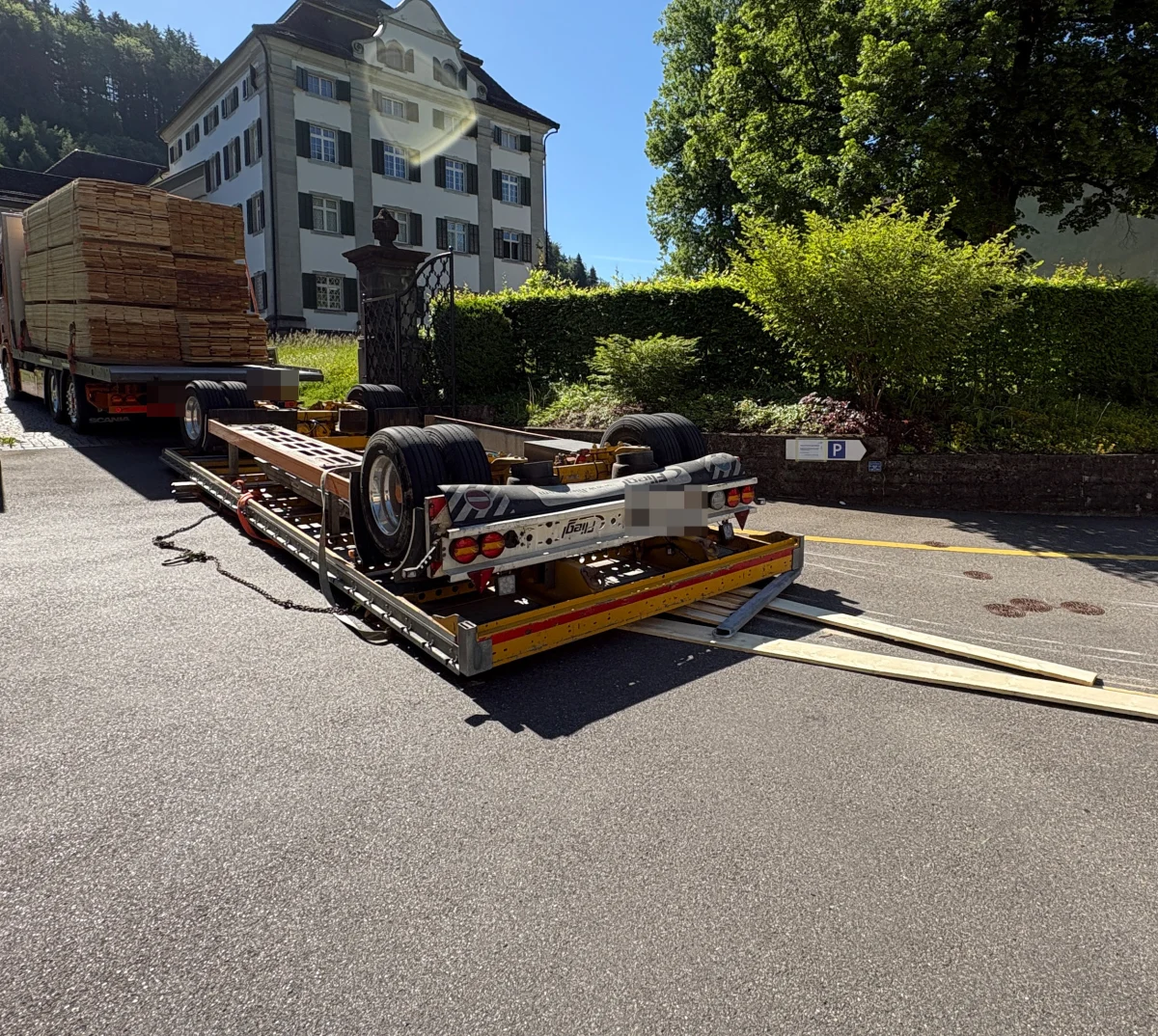 Ein Anhänger mit Holzladung liegt umgestürzt auf der Straße vor einem großen Gebäude bei sonnigem Wetter.