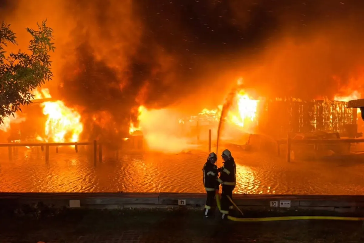 Zwei Feuerwehrleute stehen in der Nacht vor einem großen, lodernden Brand. Flammen erhellen die Umgebung.