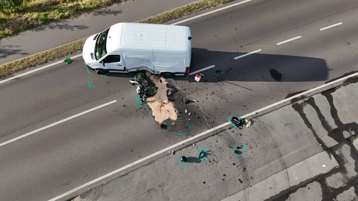 Ein weißer Van ist auf einer zweispurigen Asphaltstraße mit einem Motorrad kollidiert. Die Straße ist mit Sand und Trümmerteilen übersät. Markierungen und ein Grünstreifen sind sichtbar.