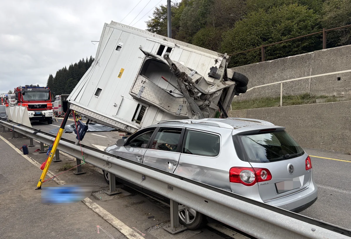 Ein umgekippter Lkw-Anhänger liegt auf einem silbernen VW Passat entlang der Autobahnleitplanke. Feuerwehrfahrzeuge sind vor Ort.