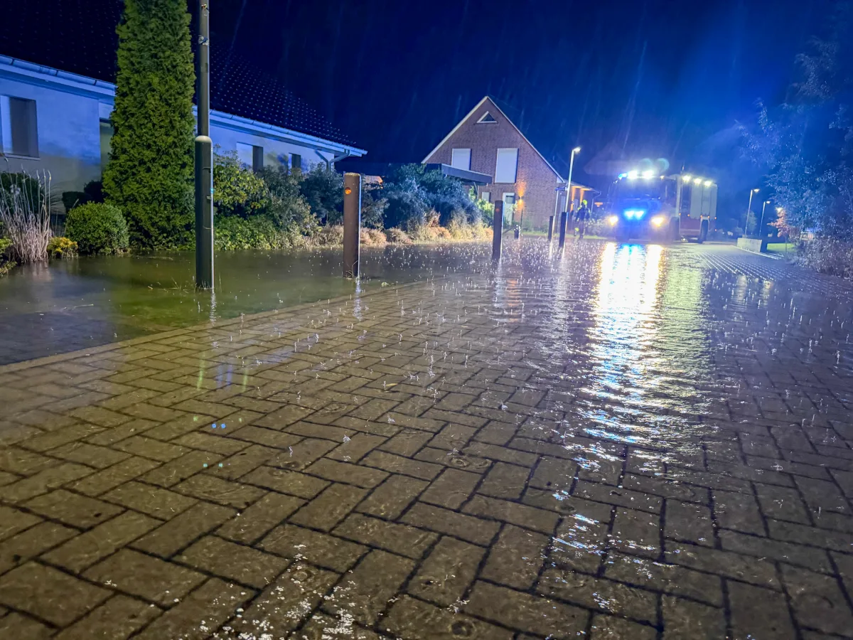 Bei Nachtbeleuchtung ist ein Feuerwehrfahrzeug vor einer überschwemmten Straße in einem Wohngebiet zu sehen. Die Straße steht unter Wasser, während Regen fällt.