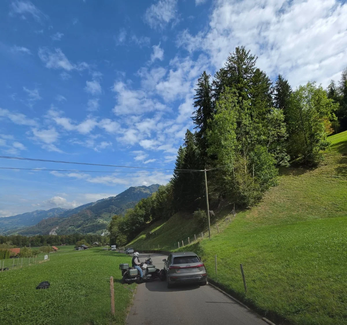 Ein Motorradfahrer fährt auf einer engen Straße durch eine grüne, hügelige Landschaft. Die Sonne scheint und der Himmel ist teilweise bewölkt.