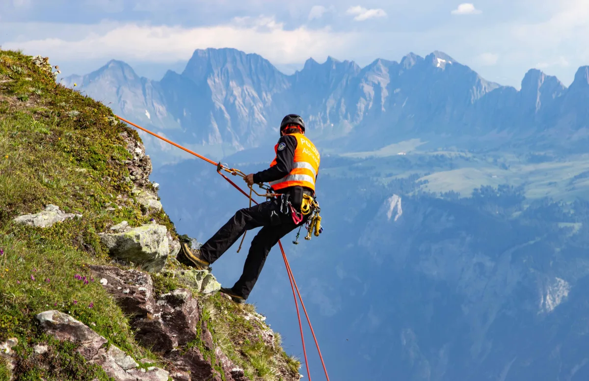 Eine Person in orangefarbener Rettungsweste und Helm seilt sich mit Seilen von einem grasbewachsenen Berghang ab. Im Hintergrund sind felsige Alpen zu sehen.