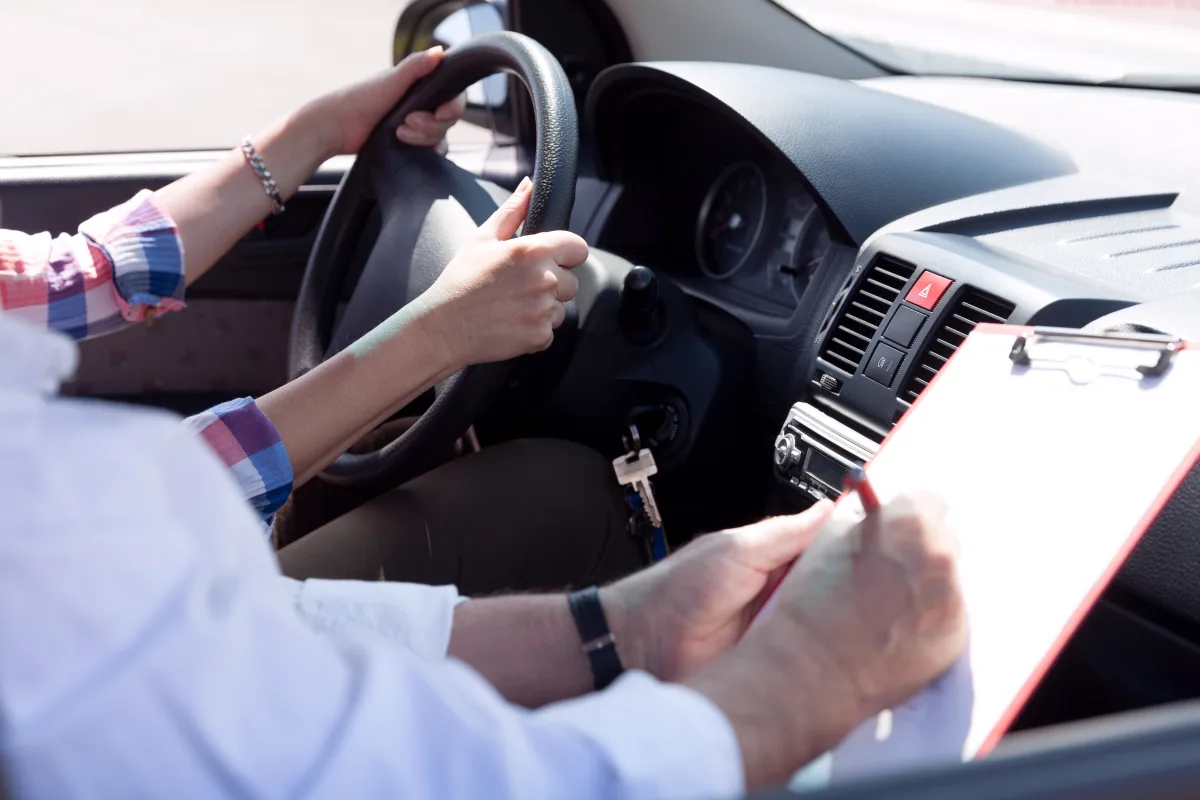 Im Auto hält eine Person das Lenkrad, während ein Fahrprüfer Notizen auf einem Klemmbrett macht. Der Innenraum des Wagens ist sichtbar.