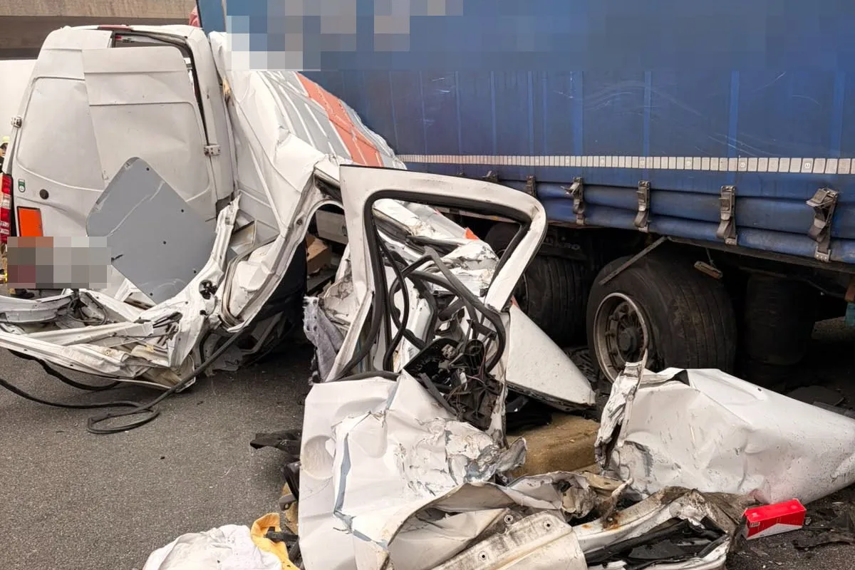 Das Bild zeigt einen stark beschädigten weißen Transporter nach einer Kollision mit einem blauen Lastwagen. Das Fahrzeug ist erheblich deformiert.