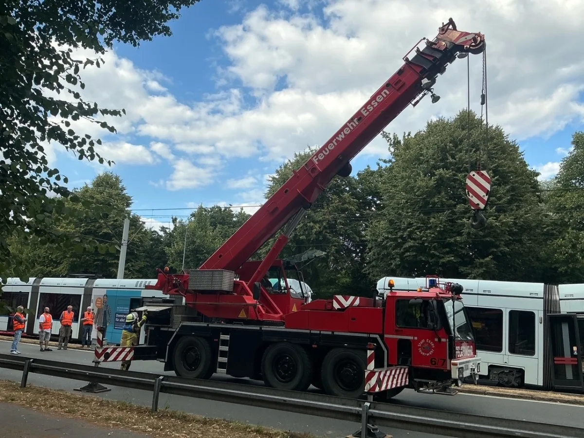 Ein großer, roter Feuerwehrkran der Feuerwehr Essen hebt eine Straßenbahn an. Mehrere Personen in orangenen Warnwesten beobachten die Szene.