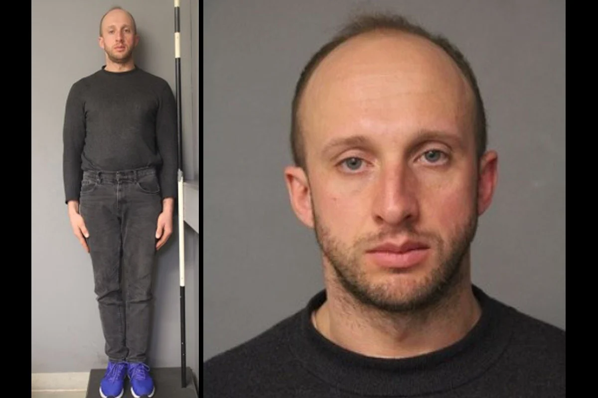 Ein Mann trägt schwarze Kleidung und steht aufrecht vor einer grauen Wand, das Bild ist in einem polizeilichen Kontext.