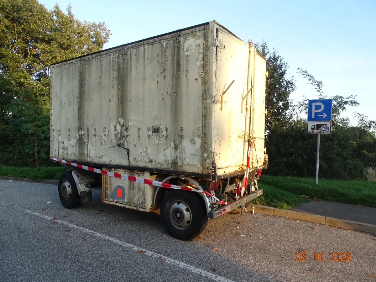Ein stark verwitterter Anhänger steht auf einem asphaltierten Parkplatz neben einem Verkehrsschild. Der Himmel ist klar.