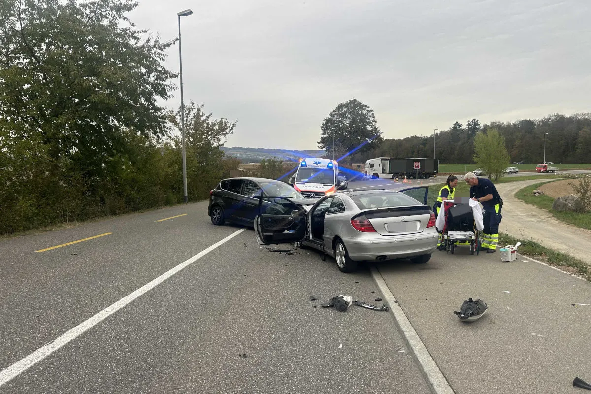 Zwei Fahrzeuge nach einem Unfall auf einer Straße, Rettungskräfte betreuen eine Person. Ein Ambulanzfahrzeug steht im Hintergrund.