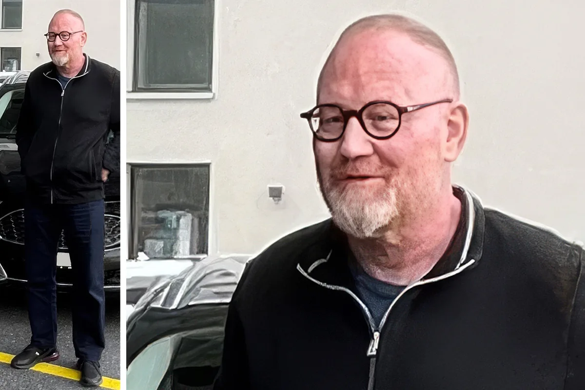Ein Mann mit Brille und hellem Bart steht neben einem schwarzen Auto vor einer weißen Gebäudewand, Fenster im Hintergrund.