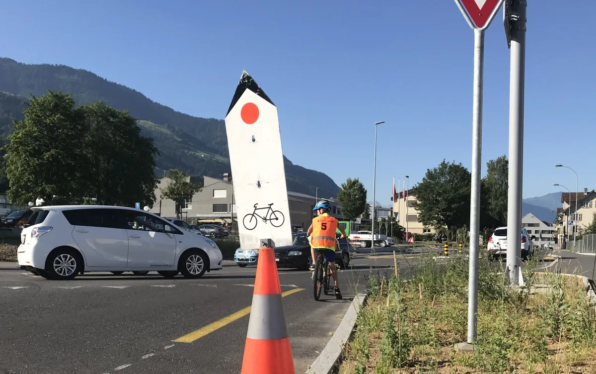 Ein Radfahrer mit Helm und Warnweste fährt an einem großen, schildförmigen Betonpfeiler vorbei, der ein Fahrrad und einen roten Punkt zeigt. Im Hintergrund sind Autos und eine ländliche Umgebung mit Bergen sichtbar.