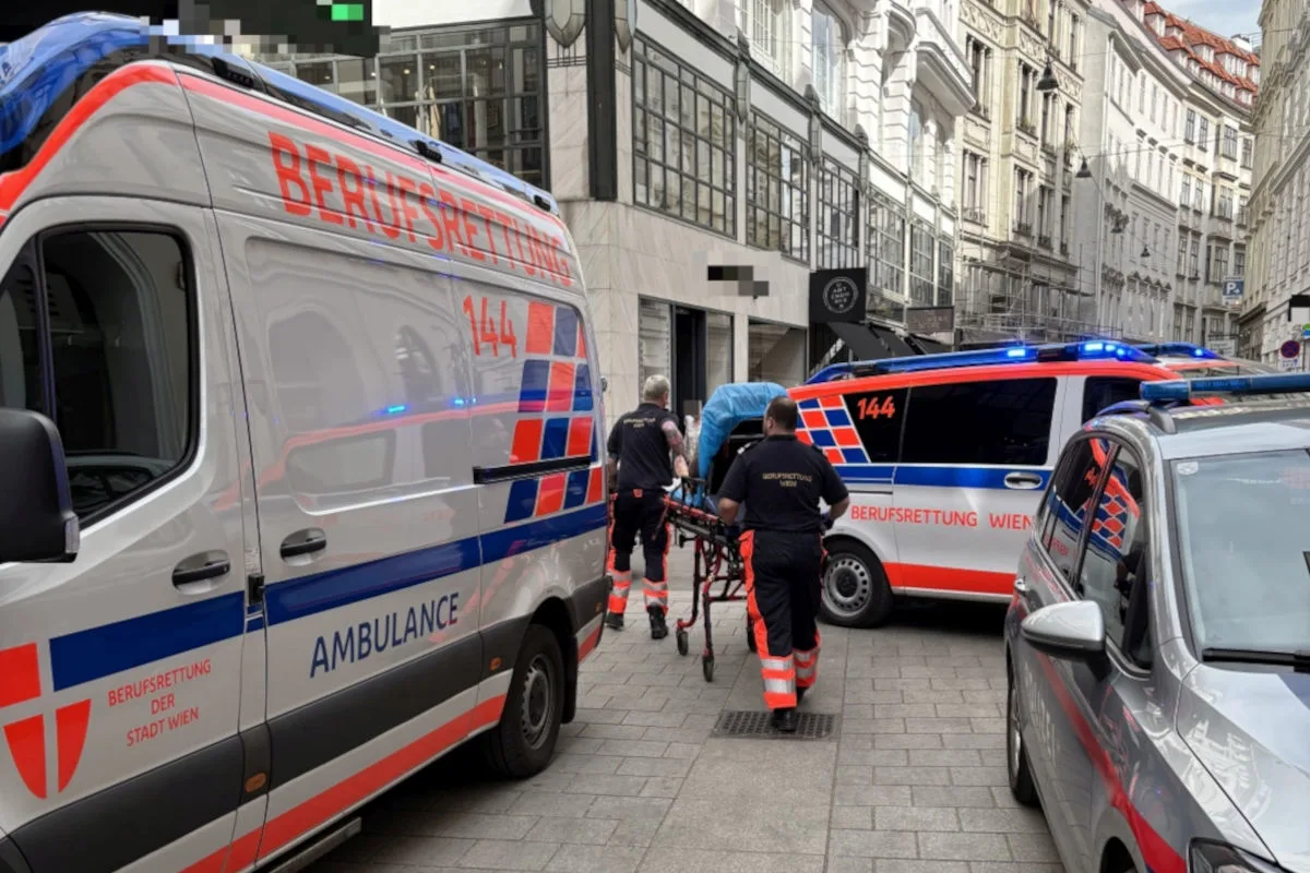 Mehrere Rettungsfahrzeuge der Berufsrettung Wien stehen auf einer Straße. Rettungskräfte schieben eine Trage in Richtung eines Krankenwagens.