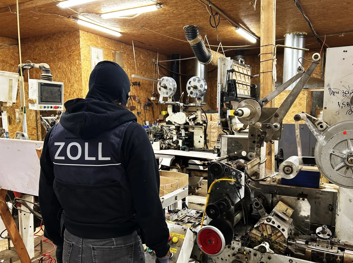 Ein Zollbeamter in dunkler Kleidung steht in einer Werkstatt voller Maschinen. Der Raum ist hell beleuchtet und wirkt industriell.