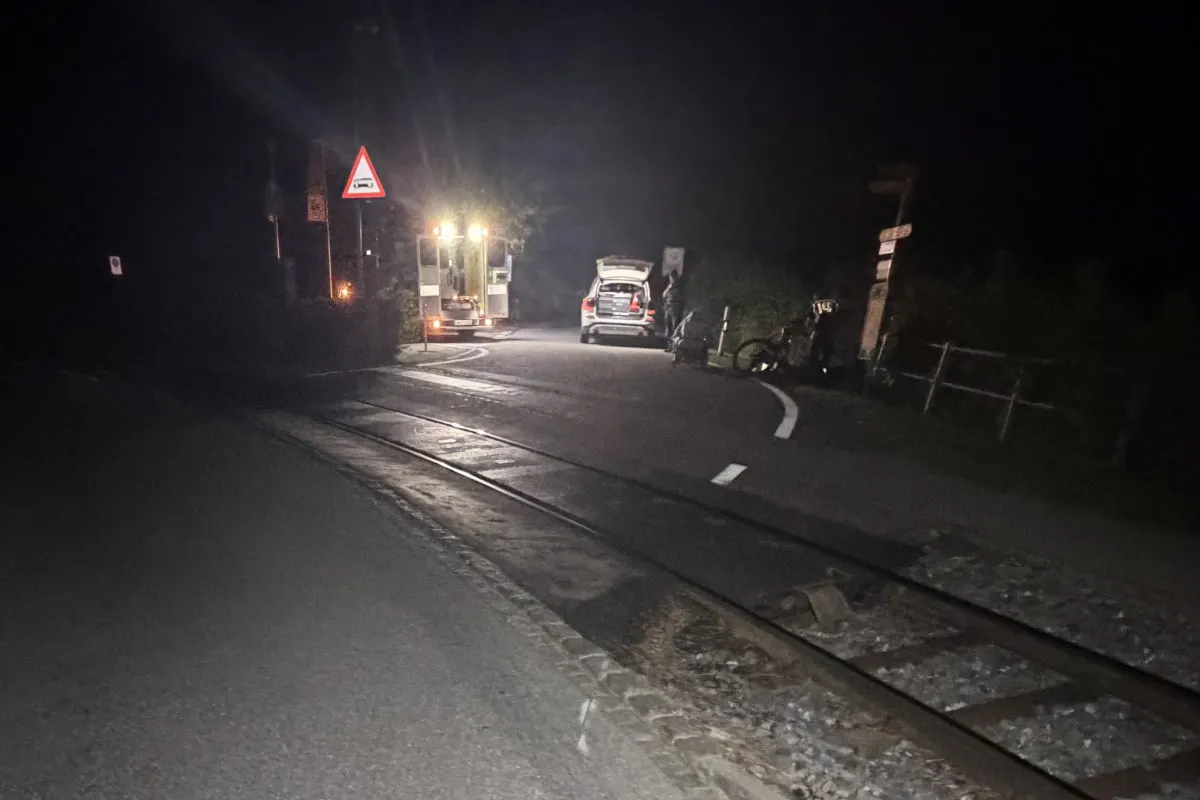 Eine dunkle Straßenkreuzung bei Nacht zeigt ein Fahrzeug mit eingeschaltetem Licht nahe einem Bahnübergang und einem Verkehrsschild.