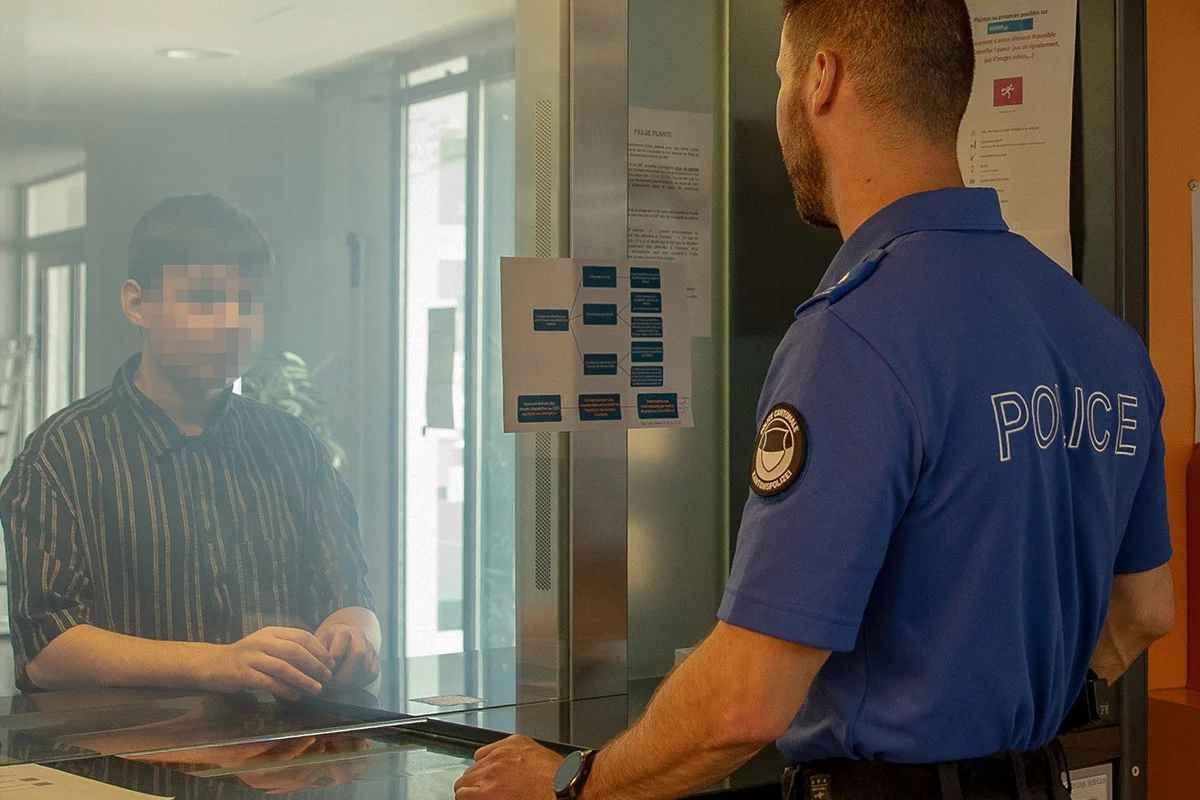Ein Polizist in blauer Uniform steht an einem Schalter und spricht mit einer Person, die durch eine Glasscheibe getrennt ist und ein gestreiftes Hemd trägt.