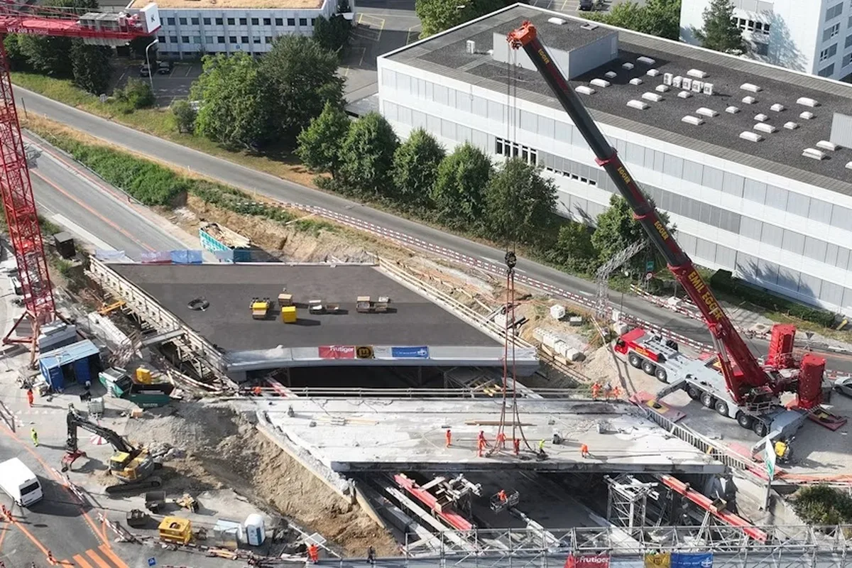 Baustelle mit zwei Kränen, Bauarbeitern in orangefarbener Kleidung und einer unvollendeten Straßenüberführung.