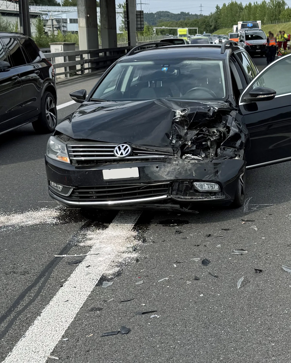 Ein schwarzer VW steht auf einer Straße, die Front ist stark eingedrückt. Im Hintergrund sind weitere Fahrzeuge und Personen zu sehen.
