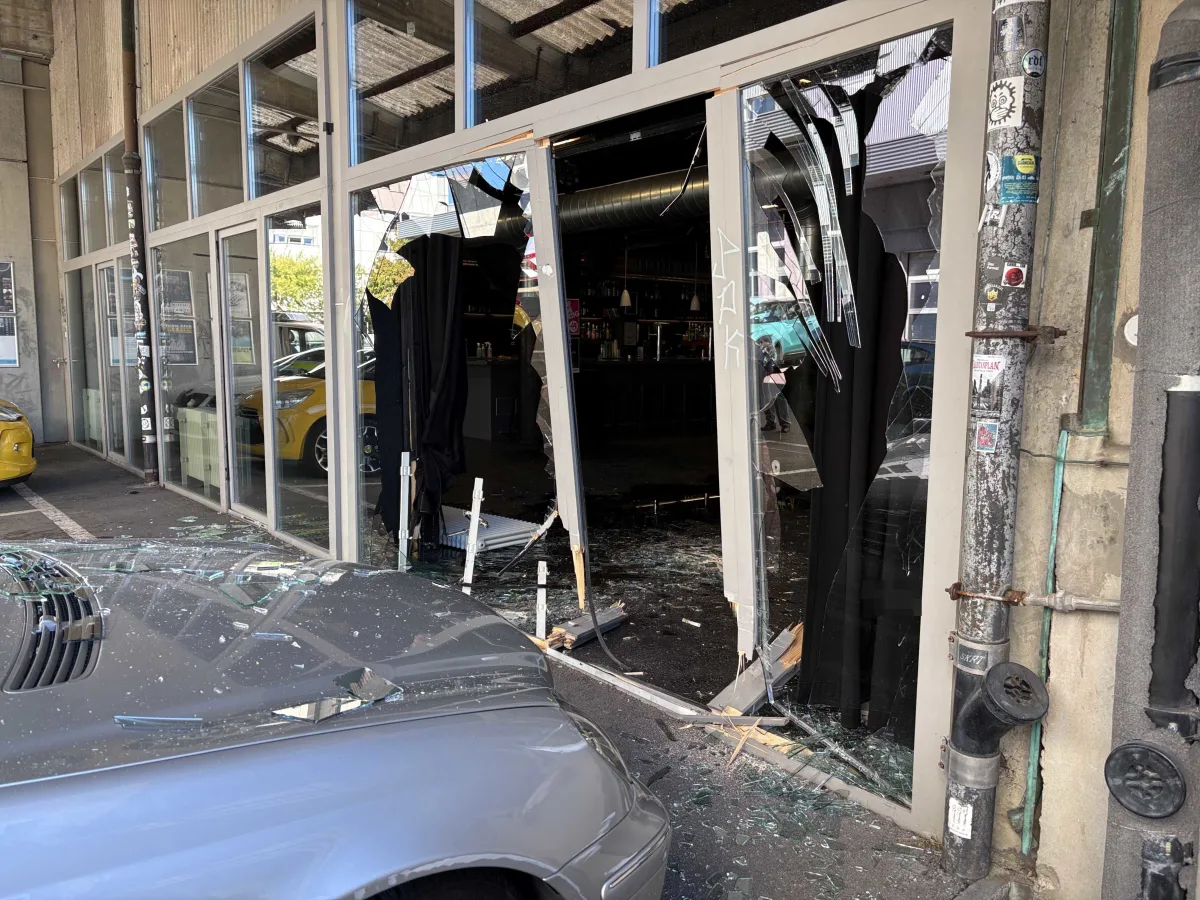 Ein silbernes Auto ist vor einer Geschäftsfassade zu sehen, deren Glastür stark beschädigt wurde. Glassplitter sind auf dem Boden verteilt.