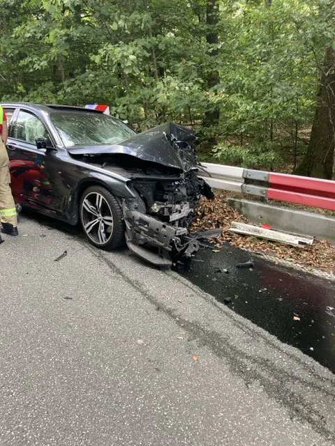 Ein schwarzes Auto steht auf einer Landstraße mit erheblichem Frontschaden an einer Leitplanke. Im Hintergrund ist Wald zu sehen.