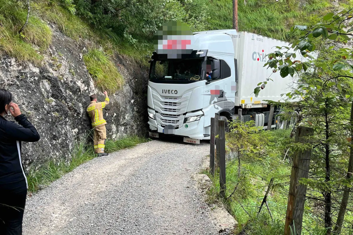 Ein weißer LKW der Marke Iveco steckt auf einem engen, unbefestigten Waldweg zwischen Felswänden und Bäumen fest. Ein Feuerwehrmann steht daneben und gibt anscheinend Anweisungen.