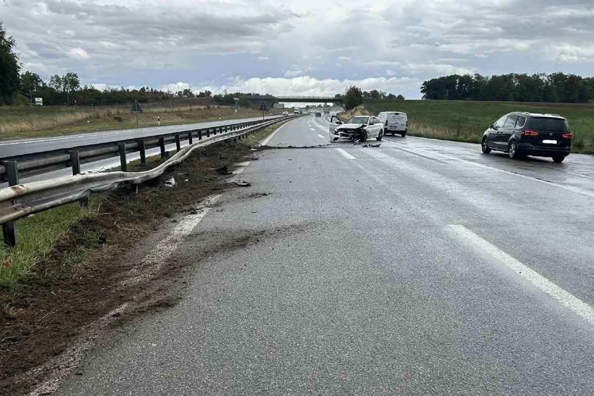 Auf der Autobahn ist ein Auto mit einer beschädigten Front zu sehen, das neben der Leitplanke steht. Die Leitplanke ist ebenfalls verformt und umgeben von Erde und Trümmern.