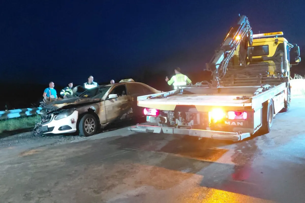 Ein beschädigtes Taxi mit deutlichen Frontschäden steht neben einem Abschleppwagen. Im Hintergrund sind Personen zu sehen, die das Geschehen beobachten.