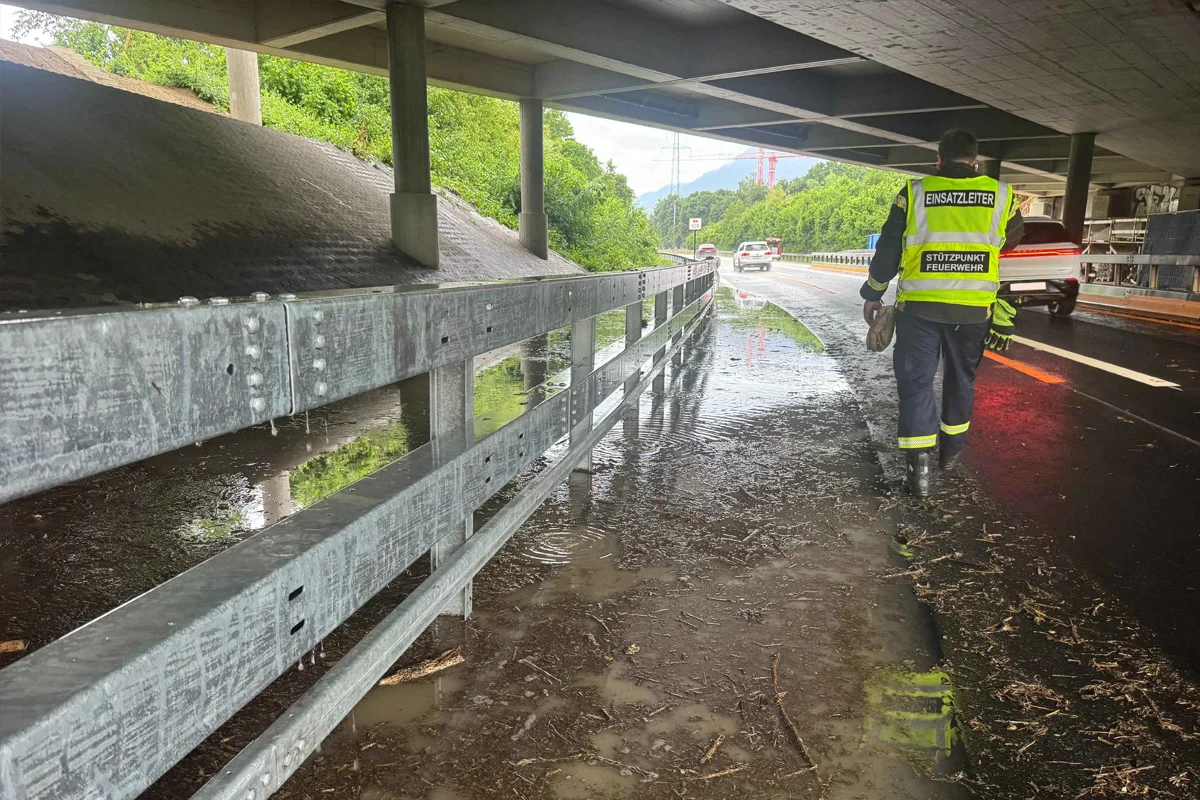 Ein Einsatzleiter der Feuerwehr, erkennbar an der reflektierenden Weste, begutachtet eine überflutete Straße unter einer Autobahnbrücke. Im Hintergrund sind Bäume und ein Fahrzeug zu sehen.