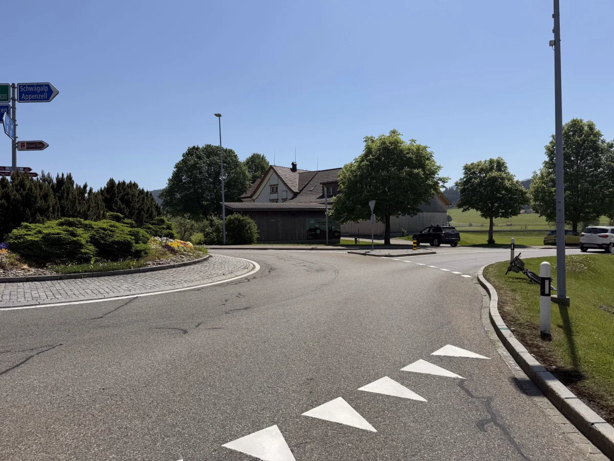 Ein Kreisverkehr in einer ländlichen Gegend mit einem Wegweiser, mehreren geparkten Autos und einem Haus im Hintergrund. Klare, sonnige Wetterbedingungen.