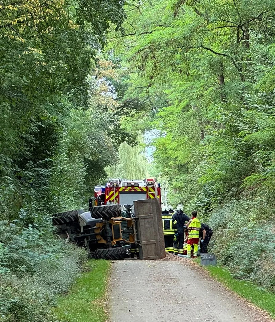 Ein Traktor liegt umgestürzt auf einem schmalen Waldweg, umgeben von Einsatzkräften in Feuerwehruniformen.