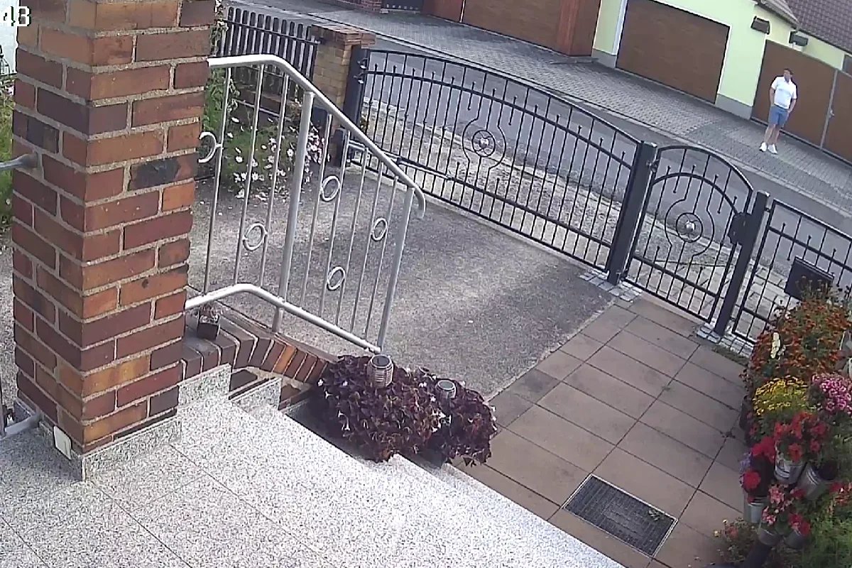 Ein Mann in kurzen Hosen und T-Shirt steht auf der Straße vor einem geschlossenen Eisentor, daneben befinden sich Blumenbeete.