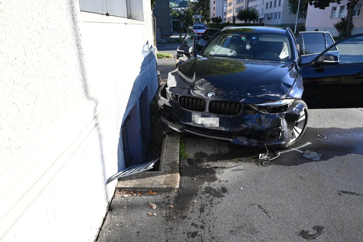 Das Bild zeigt einen schwarzen BMW mit starken Frontschäden, der gegen die Wand eines Gebäudes gefahren ist. Auf dem Boden vor dem Auto sind Trümmerteile sichtbar.
