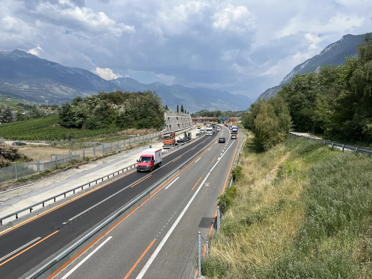 Eine vielbefahrene Autobahn führt durch eine bergige Landschaft, mit mehreren Fahrzeugen in einer Kolonne. Die Umgebung ist sonnig mit einigen Wolken am Himmel.