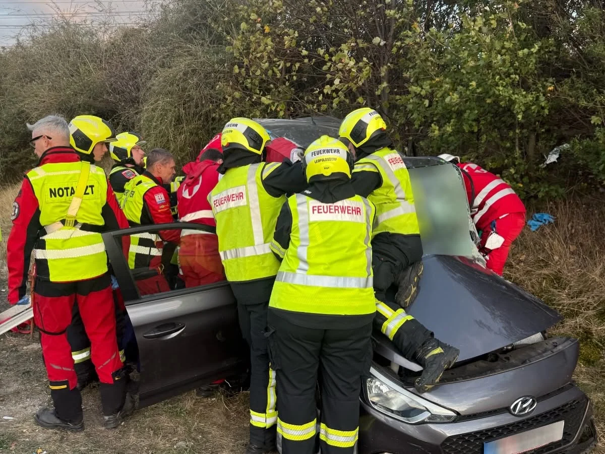 Mehrere Einsatzkräfte der Feuerwehr und Notärzte arbeiten an einem grauen Auto im Grünen, möglicherweise nach einem Unfall.