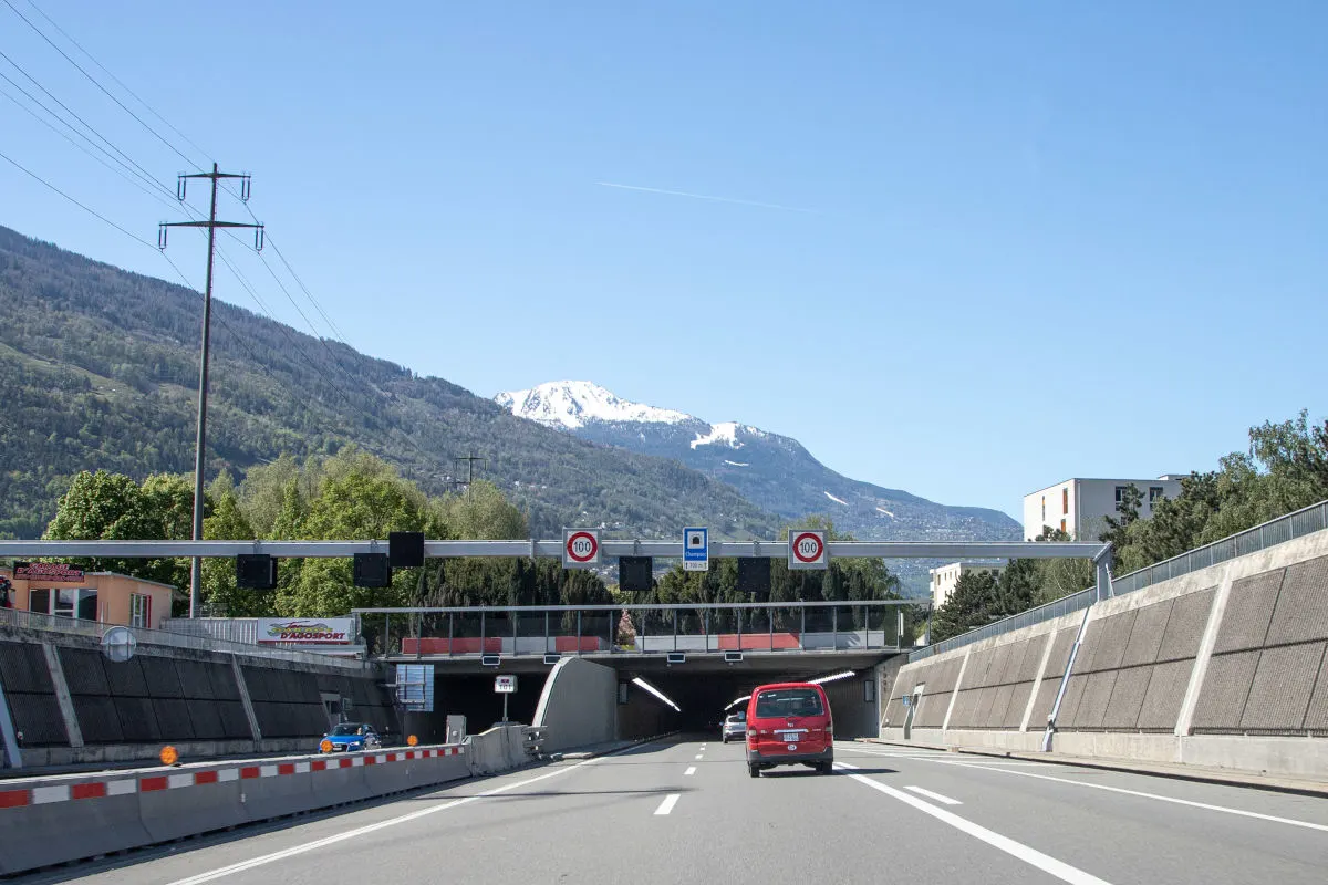 Vor einem Tunnel fährt ein rotes Auto auf einer Straße. Im Hintergrund sind bewaldete Berge mit schneebedeckten Gipfeln zu sehen.