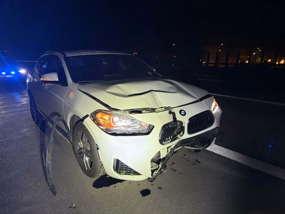 Ein weißer BMW steht mit sichtbarem Frontschaden auf einer Straße. Es ist Nacht und ein Polizeiauto ist im Hintergrund zu sehen.