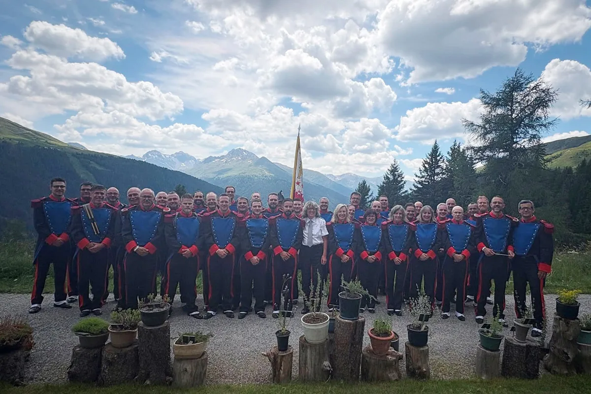 Eine Gruppe von etwa dreißig Personen trägt Uniformen und posiert vor einer pittoresken Berglandschaft mit blauem Himmel und Wolken. Im Hintergrund sind Berge und Bäume zu sehen.