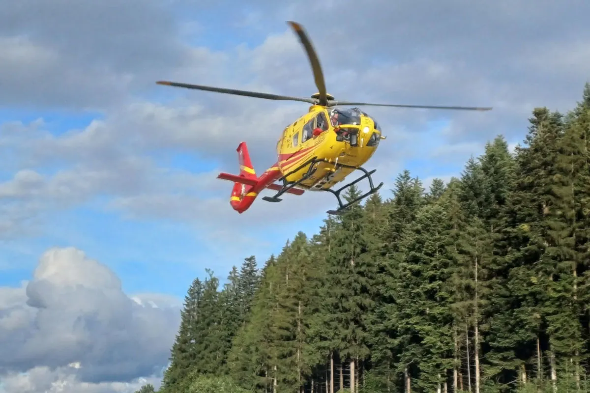 Ein gelber Rettungshubschrauber mit roten Markierungen schwebt über einem dichten Wald. Der Himmel ist leicht bewölkt und in der Ferne zu sehen.