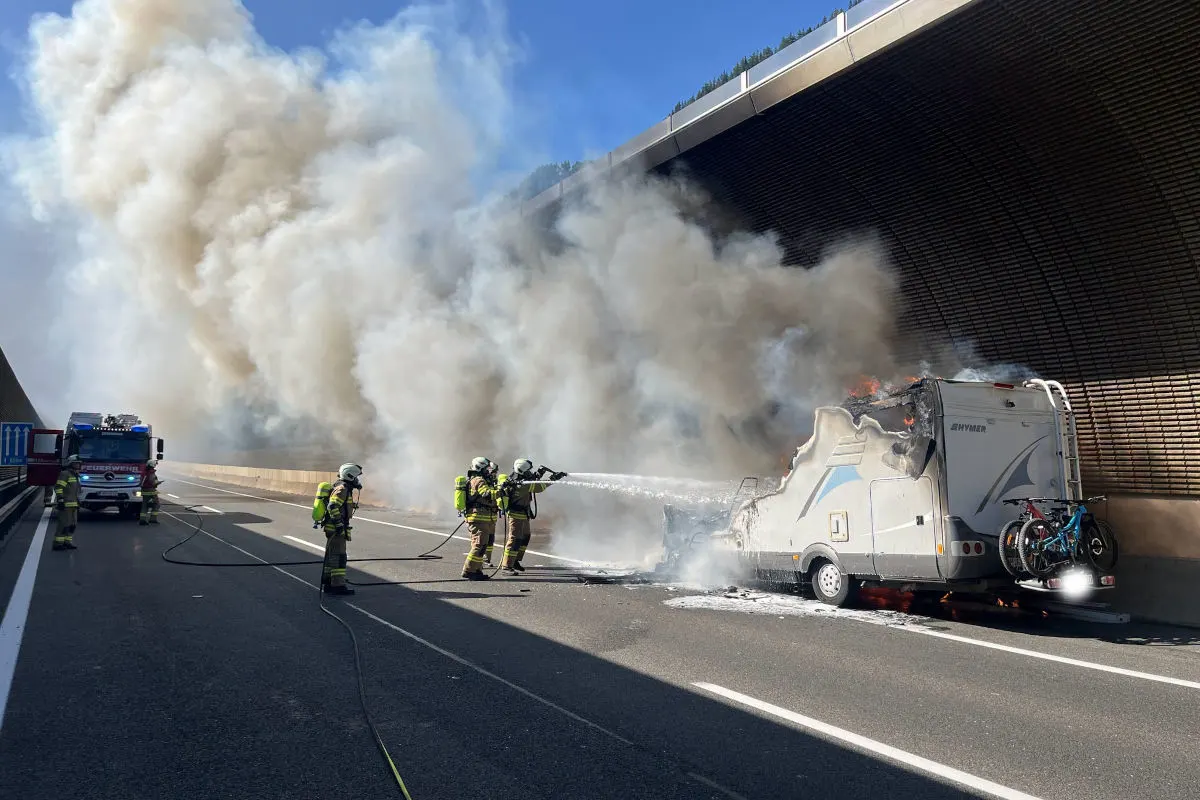 Auf der Autobahn löschen Feuerwehrleute in voller Montur mit Schläuchen ein brennendes Wohnmobil, während dicke Rauchwolken aufsteigen.