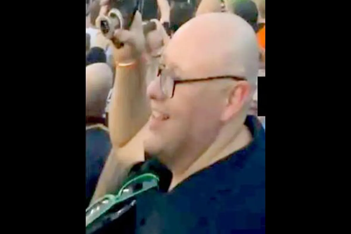 Ein Mann mit Glatze und Brille lächelt, während er in einer Menschenmenge steht und fotografiert. Er hält eine Kamera hoch.