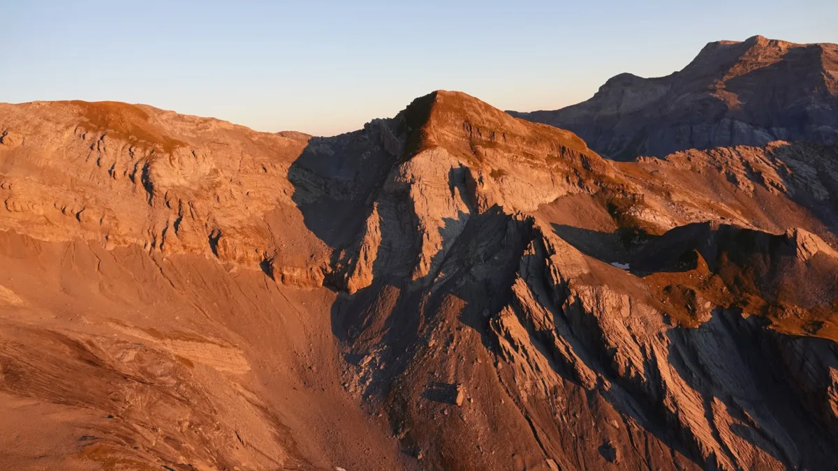 Ein Bergmassiv wird bei Sonnenuntergang beleuchtet, wodurch Schatten die Felsformationen betonen. Die Szene zeigt eine raue, felsige Landschaft ohne sichtbare Menschen oder Fahrzeuge.
