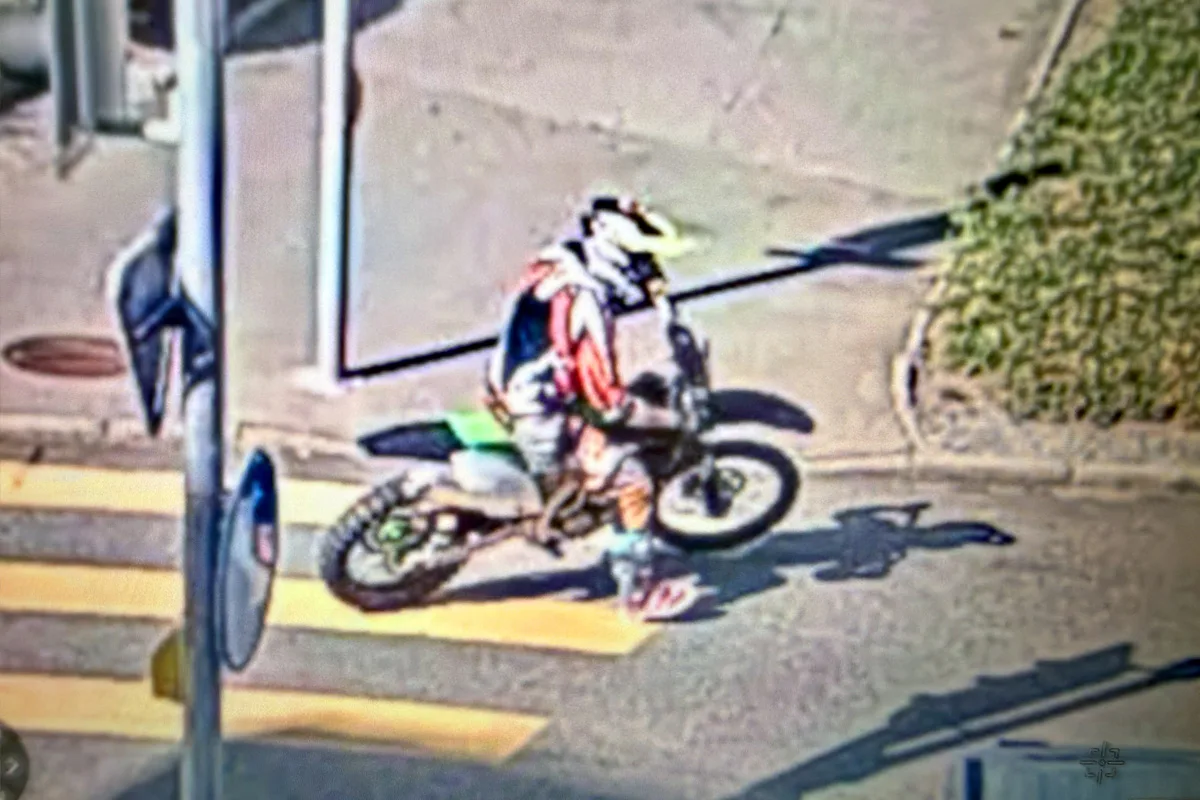Ein Motorradfahrer mit Helm fährt bei Tag auf einem Zebrastreifen in einer städtischen Umgebung. Die Straße und Verkehrszeichen sind sichtbar.
