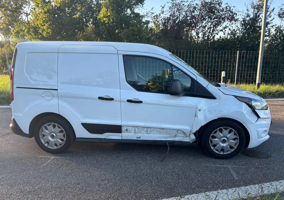 Ein weißer Lieferwagen mit Seitenschaden steht auf einer asphaltierten Straße. Im Hintergrund sind Bäume und ein Zaun sichtbar.