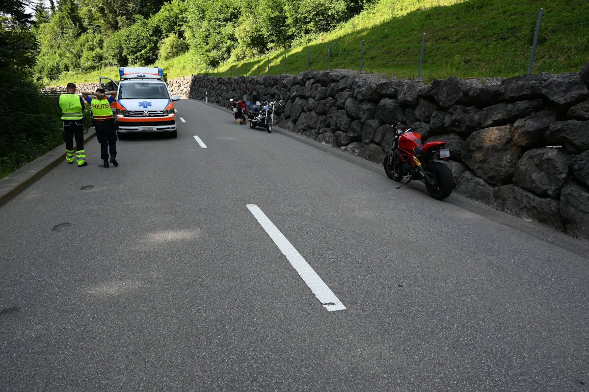 Auf einer Landstraße stehen ein Krankenwagen und mehrere Motorräder. Zwei Personen in uniformähnlicher Kleidung stehen links.