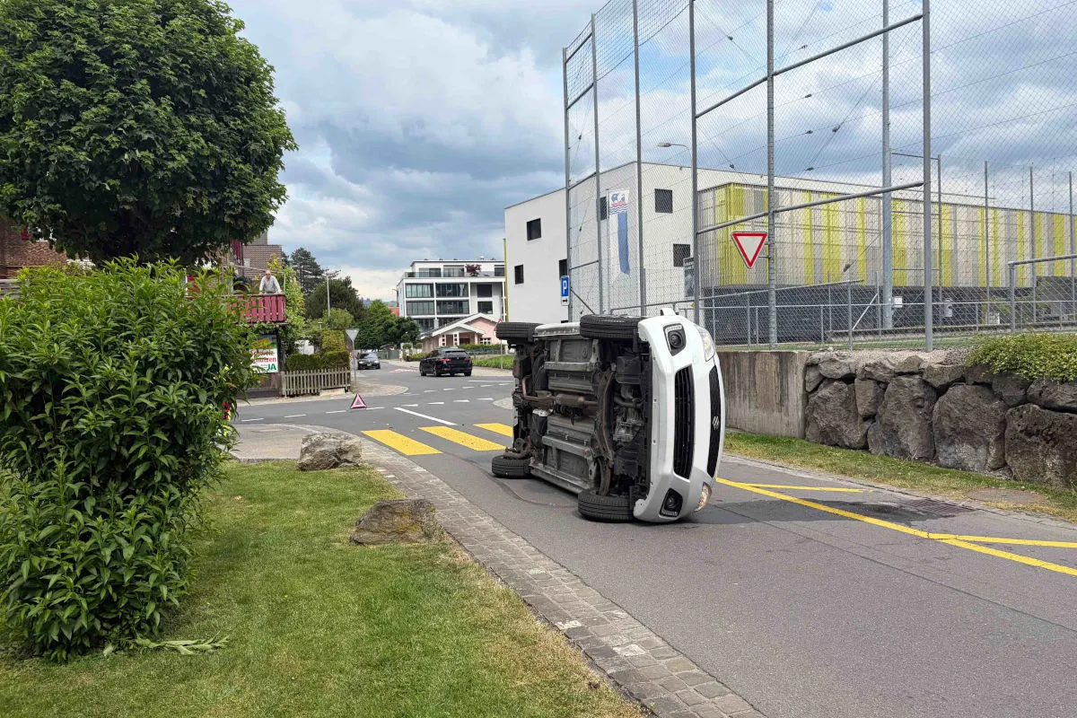 Ein weißes Auto liegt auf der Seite an einer Straßenkurve, umgeben von Wohnhäusern und einer Parkanlage.
