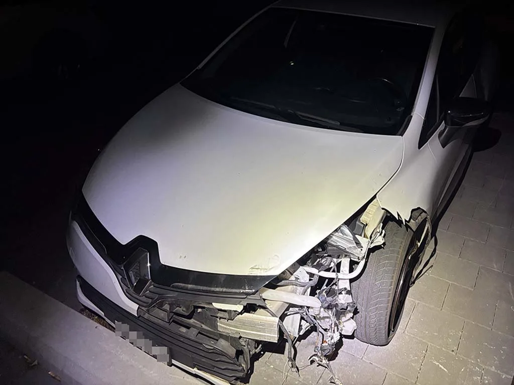Ein stark beschädigtes weißes Auto ist bei Nacht zu sehen. Die Front des Fahrzeugs weist erhebliche Schäden auf, insbesondere an der rechten Ecke.